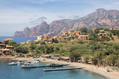 Village de Girolata vu depuis la baie, avec ses maisons colorées au bord de l'eau et les falaises rouges caractéristiques de la réserve de Scandola en arrière-plan.